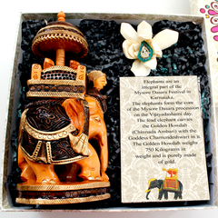 A carved wooden elephant sculpture in a festive attire, seated on a black base, with a white flower and a green jewel adornment next to it, all presented in a box with a description card.