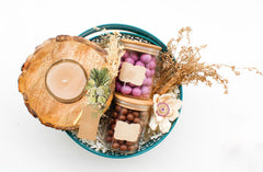 A festive gift basket arrangement with glass canisters, a rustic bark slice tea light holder with a candle, and decorative elements, presented in a circular basket.
