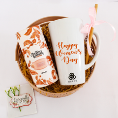 A custom-printed white latte mug with orange text reading 'Happy Women's Day' from a brand called BRAND, along with Maverick coffee, placed in a wicker basket with a view of a gift box and a card with the phrase 'Specially for you'.