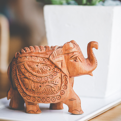 A wooden elephant figurine with intricate carvings, placed on a white surface, with a blurred background.