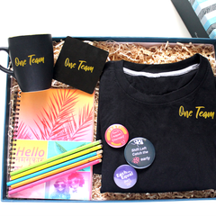 A gift set containing a black t-shirt with 'One Team' text, a spiral hardbound diary, eco-friendly paper pencils, a ceramic coffee mug with coaster, and three button badges, all displayed in a customised keepsake box.