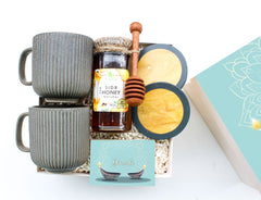 A collection of ceramic coffee mugs and coasters in a gift basket, with a jar of honey, coconut shells, and a greeting card.