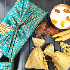 A green gift box with a gold-colored lid, adorned with a golden-brown leaf-like pattern and tied with a yellow ribbon. The box is presented on a dark background alongside some decorative leaves and a candle.