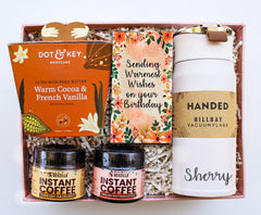 A gift box containing a moisturizer, gourmet coffee in jars, a body butter tube, a vacuum flask, and a greeting card.
