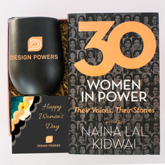 A black gift box with a sleeve, a book titled 'WOMEN IN POWER' with a subtitle 'THEIR VOICES, THEIR STORIES', and a black mug with 'DESIGN POWERS' logo, all arranged on a surface. The box has a pattern of women's faces, and there is a personalized greeting card with a floral design.