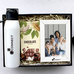 A corporate gift box containing a white sipper bottle, a chocolate bar, and a picture frame with a welcoming message and a picture of a family.