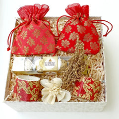 A gift hamper containing two red and gold floral patterned pouches, a set of Ferrero Rocher chocolates, and a set of white designer candles with gold accents, all arranged in a white designer-branded gift box.