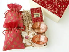 A gift hamper containing copper diyas and 2 pouches with almonds and biscotti, all packaged in a designer-branded gift box with a 'Happy Diwali' card.
