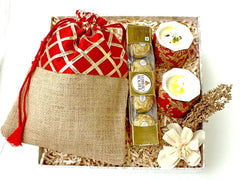 A designer-branded gift box containing a pouch of trail mix, a set of Ferrero Rocher chocolates, and a set of designer candles, all adorned with decorative elements.